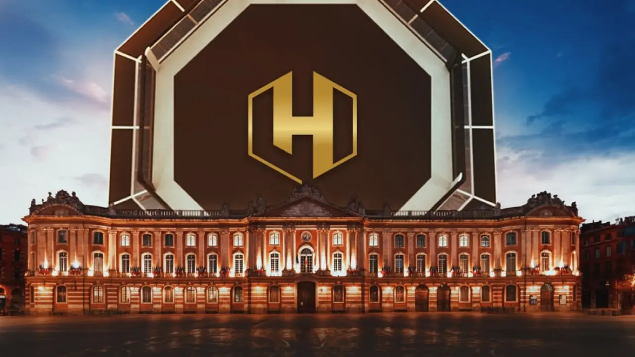Un imposant bâtiment historique aux fenêtres chaleureusement illuminées au crépuscule, surmonté d'un grand hexagone moderne orné d'un audacieux logo géométrique doré en forme de « H ». Le ciel est d'un bleu doux.