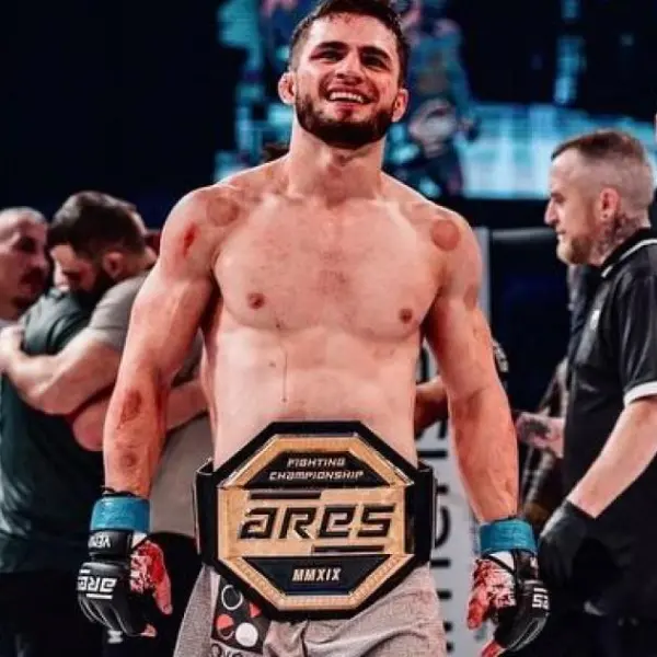 Un combattant de MMA victorieux, souriant, porte une ceinture de champion siglée « Ares ». Il est sur un ring, avec des spectateurs en arrière-plan qui célèbrent. L'ambiance est à la victoire.