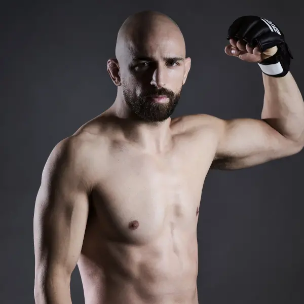 Un homme musclé, chauve et barbu pose avec assurance, le bras droit fléchi. Il porte un gant noir. Le fond est d'un gris foncé uni.