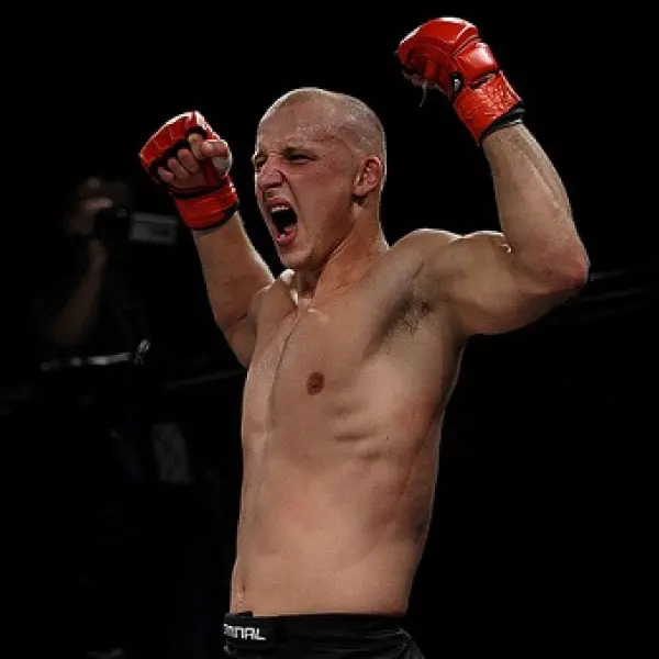 Un combattant de MMA torse nu avec des gants rouges lève les deux bras et crie de joie dans une arène faiblement éclairée.
