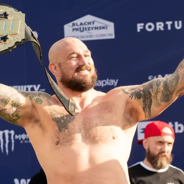 Combattant de MMA barbu et tatoué, torse nu, levant les bras en souriant avec une ceinture de champion sur l’épaule, lors d’une cérémonie en extérieur avec fond promotionnel KSW.