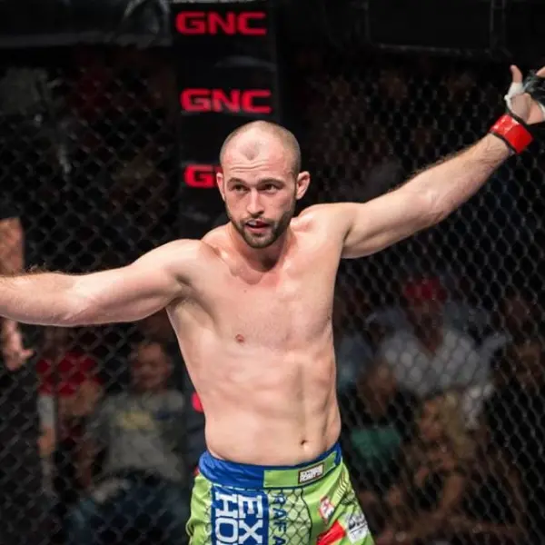 Combattant de MMA torse nu dans une cage, levant les bras en signe de victoire, portant un short de combat coloré, devant une foule en arrière-plan.