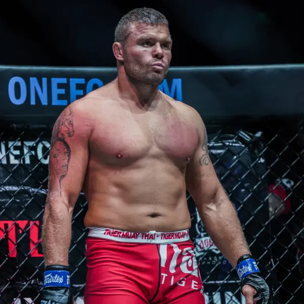Un combattant musclé, vêtu d'un short rouge et de gants bleus, se tient concentré dans une cage de MMA. En arrière-plan, on aperçoit une partie du logo du ONE FC. L'ambiance est intense.