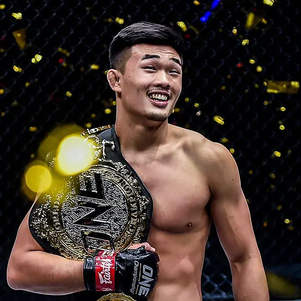 Combattant de MMA torse nu aux cheveux courts souriant largement, tenant une grande ceinture de championnat dans une arène en cage ornée de confettis dorés, exsudant la victoire et la joie.