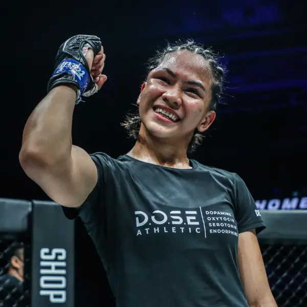Une athlète souriant, vêtu d'un maillot noir et de gants bleus, lève le poing en signe de victoire dans une cage de MMA. Ambiance énergique et triomphante.