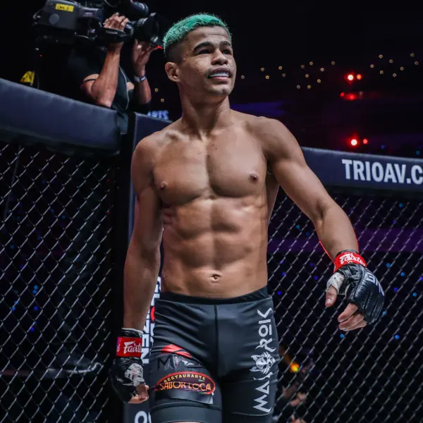 Un combattant de MMA aux cheveux verts se tient avec assurance dans une cage, vêtu d'un short et de gants noirs. La caméra capture son expression déterminée sous une lumière vive.