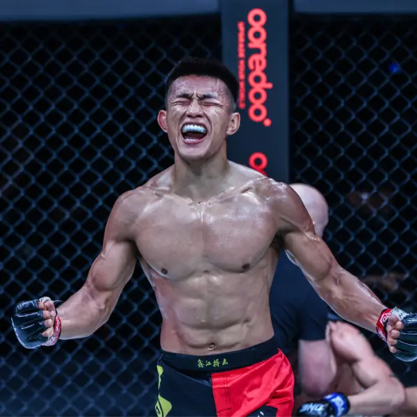 Un combattant musclé en short noir et rouge crie triomphalement les yeux fermés à l'intérieur d'une cage, transmettant intensité et victoire.