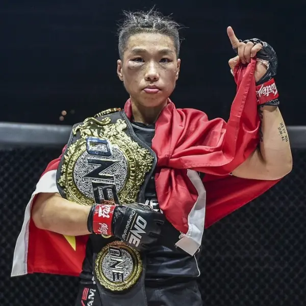 Artiste martial portant une ceinture de champion et un drapeau rouge. Il lève un doigt, l'air confiant, dans une arène faiblement éclairée, exprimant victoire et fierté.