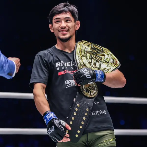 Un homme aux cheveux noirs et courts sourit et porte une ceinture de champion d'or sur son épaule. Il porte un t-shirt noir et des gants de MMA bleus sur un ring de boxe.